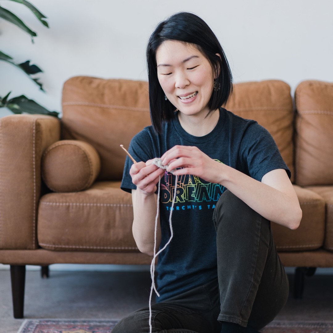 Picture of April Lee looking down and crocheting while sitting on the ground in front of a brown couch