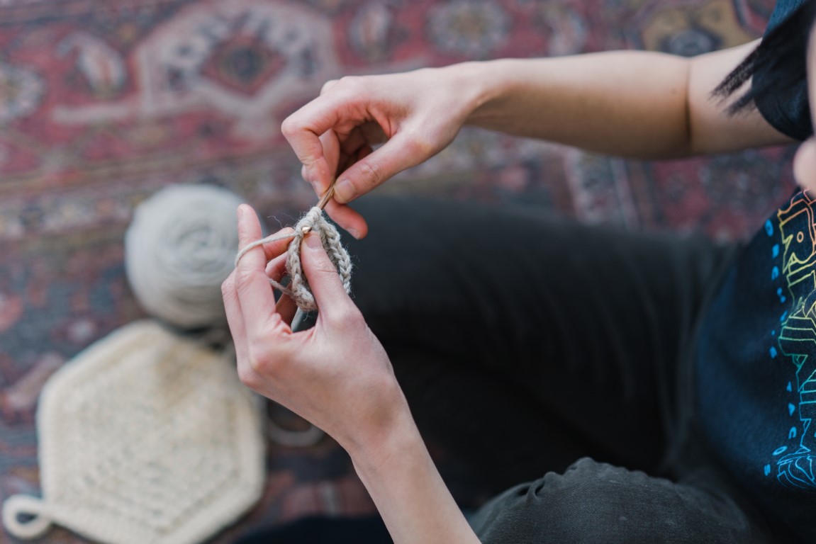 Picture of April's hands crocheting while sitting on the ground with a hexagon crochet potholder in the background